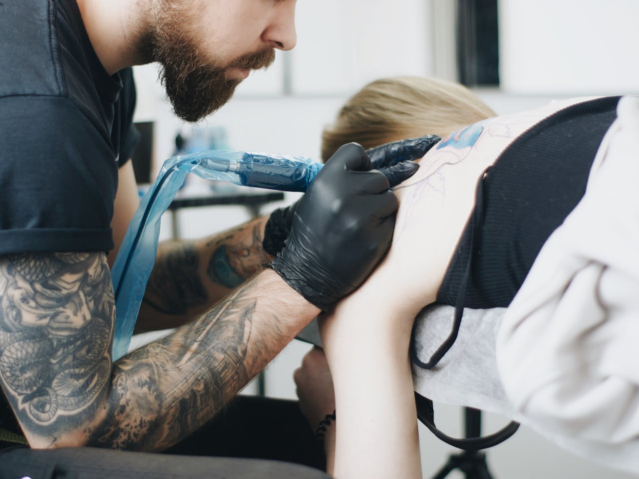 Tattoo artist with beard and gloves creating a back tattoo in a studio setting.